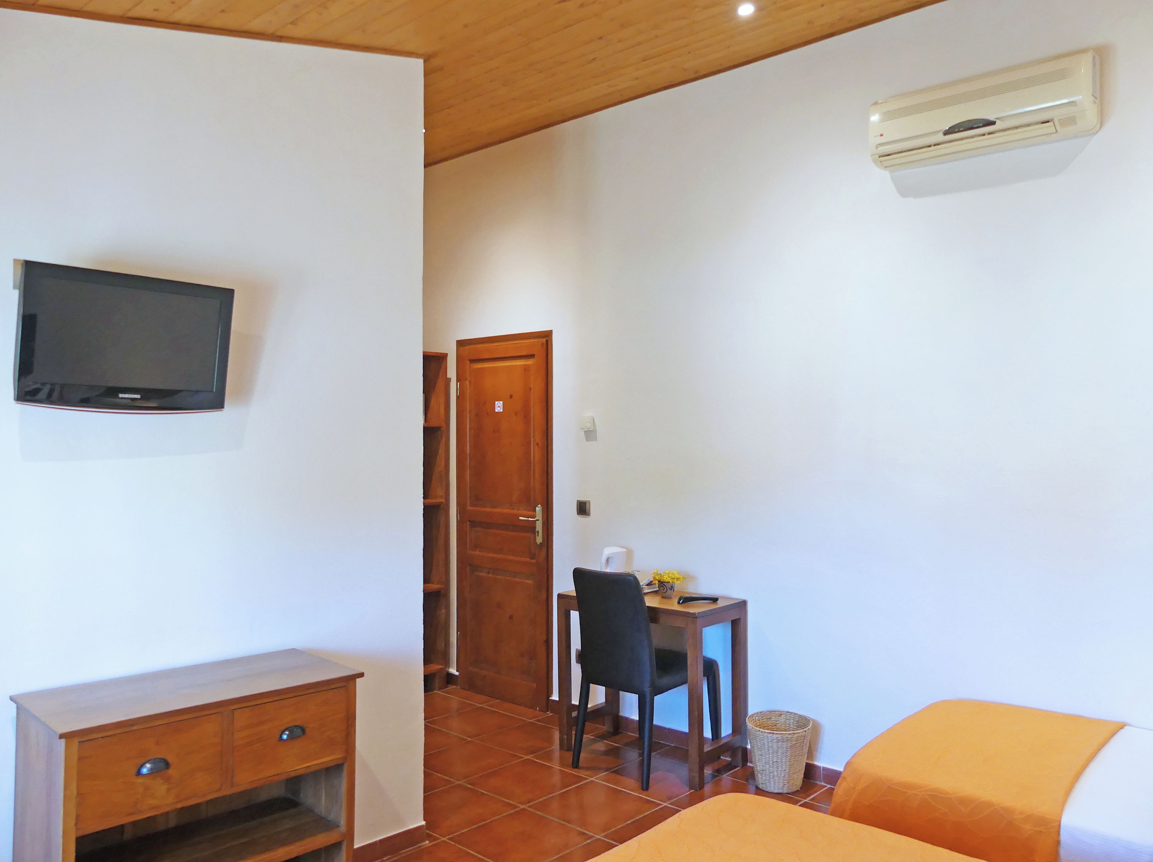 Ein Zimmer mit an der Wand montiertem Fernseher, einem Holztisch mit Stuhl, einem kleinen Tisch ebenfalls mit Stuhl und einer Klimaanlage an der Wand.