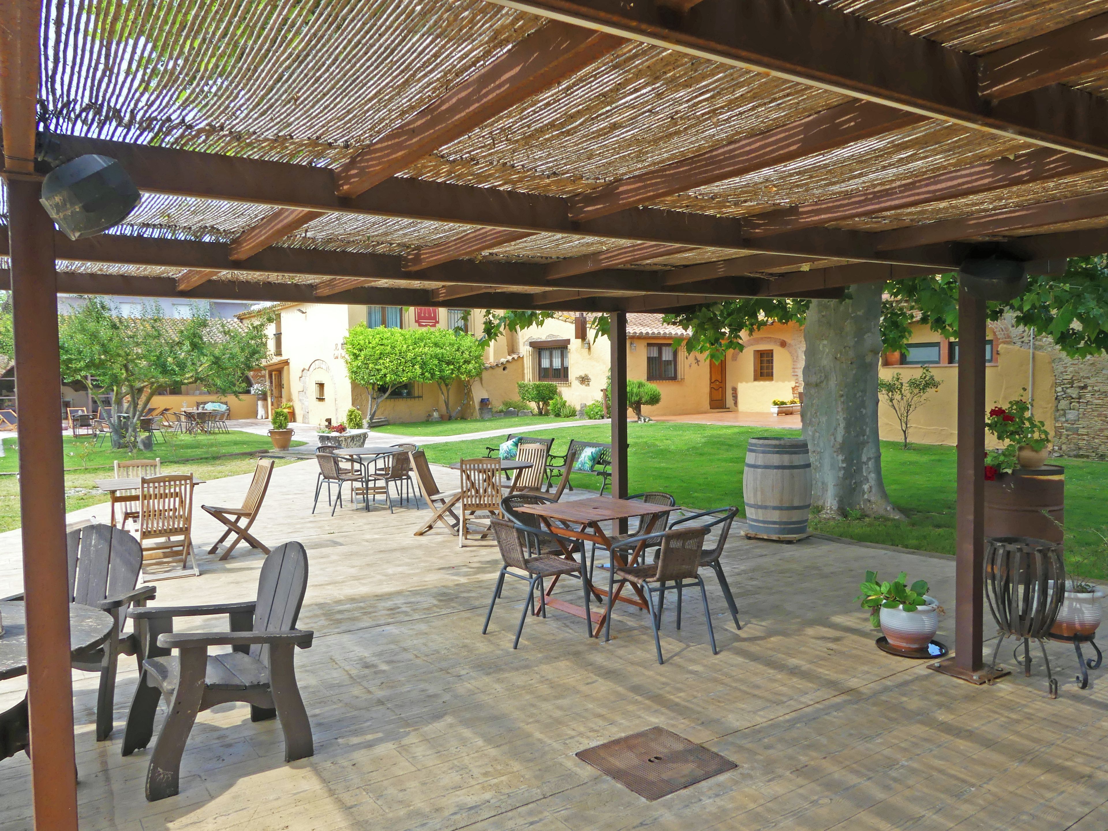 Un patio al aire libre con mesas y sillas, una pérgola en lo alto y vista a un edificio con muro de piedra al fondo.