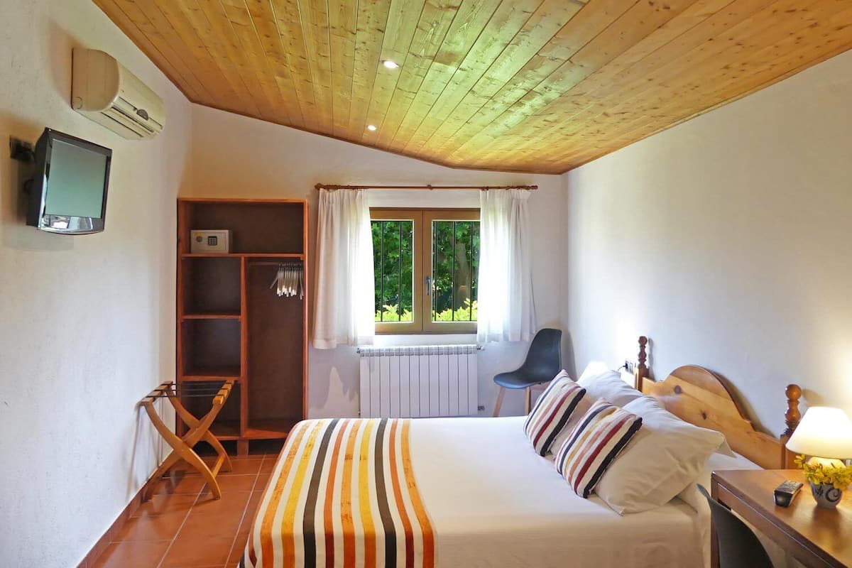 Une chambre avec un plafond en bois, un lit avec une literie à rayures, une fenêtre avec des rideaux, un radiateur, un téléviseur fixé au mur et une petite table avec une lampe et des fleurs.