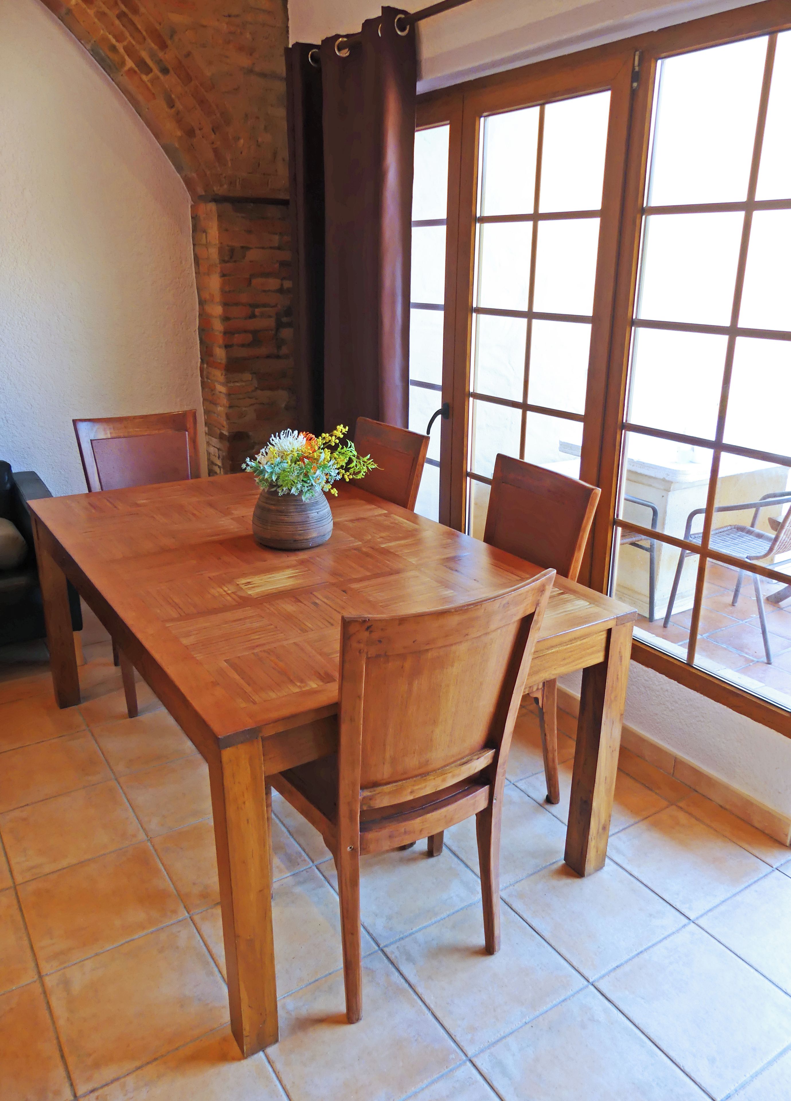 Una mesa de madera con sillas, un jarrón con flores, un muro de ladrillo y un ventanal con vistas.