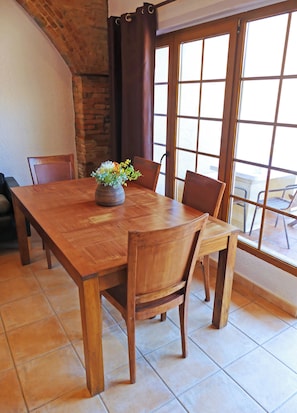 Een houten eettafel met stoelen, een vaas met bloemen, een bakstenen muur en een groot raam met uitzicht.