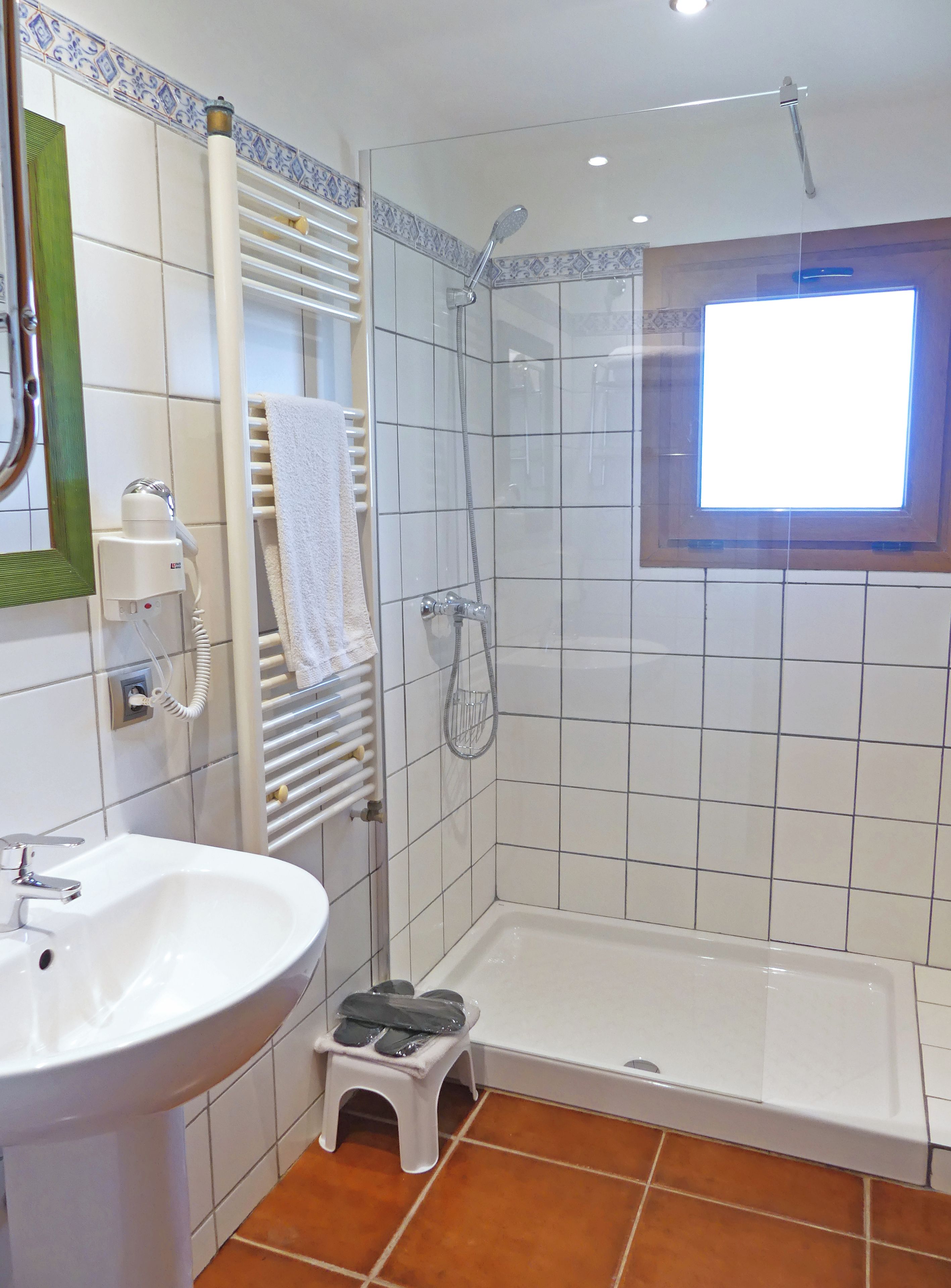 Un baño con lavamanos blanco, un taburete pequeño, ducha con puerta de cristal y una ventana.