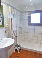 Un baño con lavamanos blanco, un taburete pequeño, ducha con puerta de cristal y una ventana.