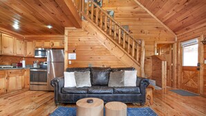 A log cabin interior with a wooden staircase, a leather sofa, a dining table, and a kitchen area. - Blue Mist Lodge: Hot Tub, Outdoor Lounge, MORE (Sevierville)