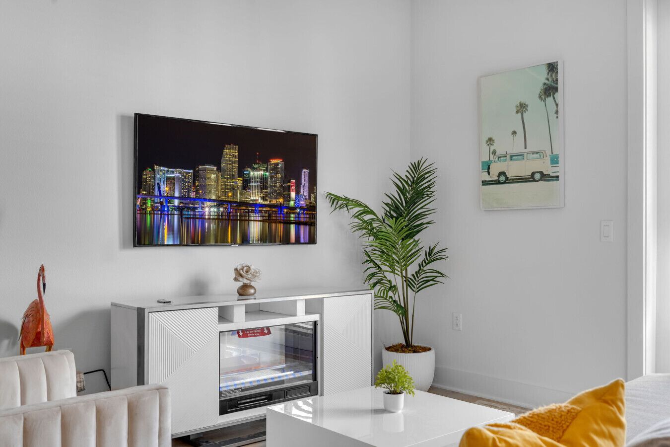 Uma sala de estar moderna com uma televisão de tela plana grande exibindo um horizonte urbano, um móvel branco para eletrônicos, uma planta em vaso e uma almofada amarela em um sofá branco.
