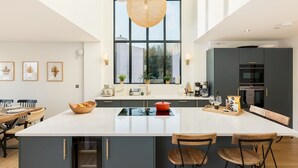 A modern kitchen with a large island, dark cabinetry, and a view of the outdoors through large windows.