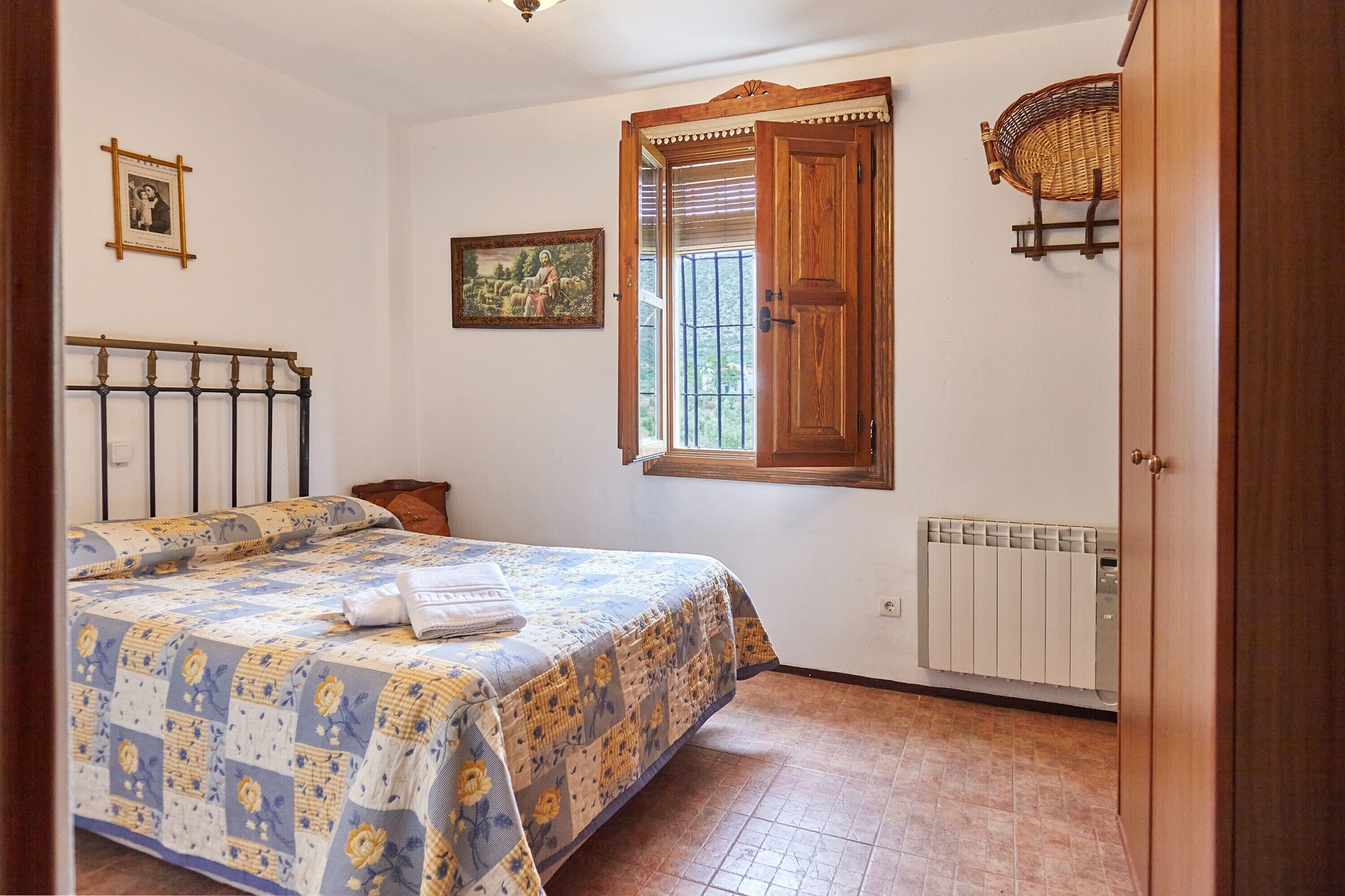 Un dormitorio con cama, ventana con contraventanas de madera, radiador y una cesta sujeta a la pared.