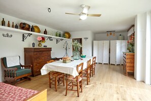 Uma sala de jantar com mesa posta para seis pessoas, cadeiras de madeira, um ventilador de teto e uma cozinha ao fundo.