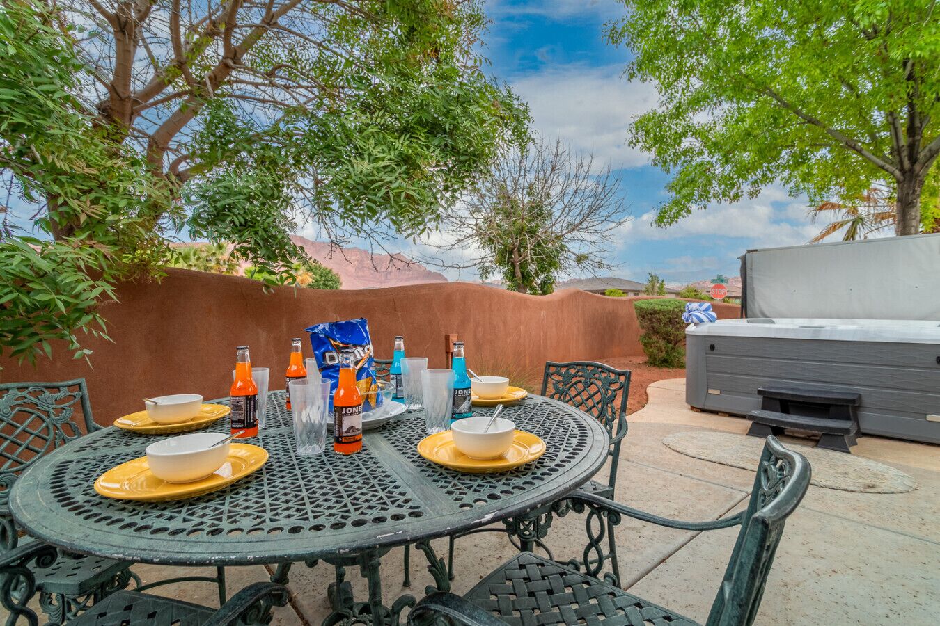 An outdoor dining area with a round table set for four, surrounded by chairs. There is a grill and a bag of chips on the table.
