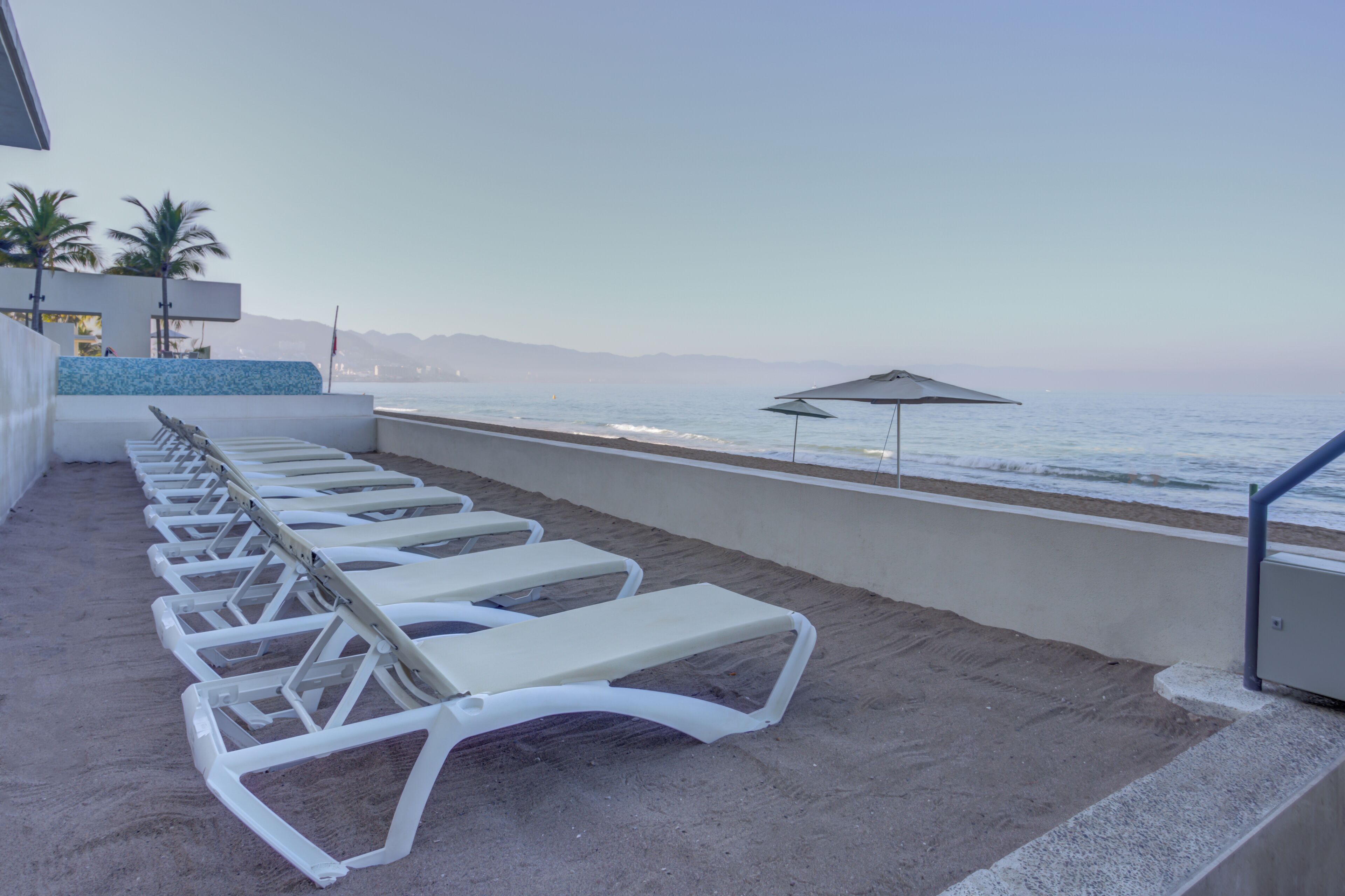 Une rangée de chaises longues blanches, protégées par des parasols, est disposée sur une terrasse en béton donnant sur la plage.