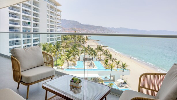 A balcony with wicker furniture, a glass table, and a view of a beach and pool area.
