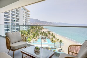 Un balcón con mobiliario de mimbre, una mesa de vidrio y vista a la playa y la piscina.