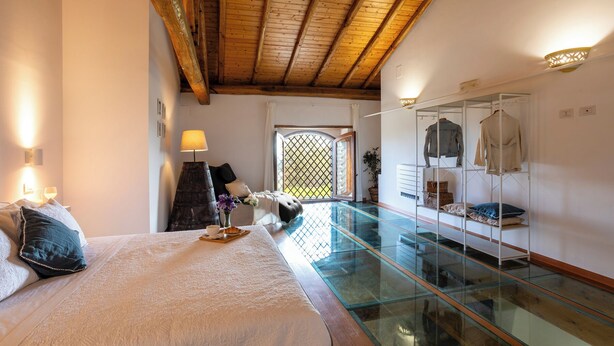 A bedroom with a glass floor, wooden ceiling beams, a bed with pillows, a nightstand with a lamp, and a wardrobe with clothes.