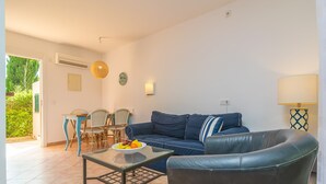 Un salon avec un canapé bleu, une table basse en verre sur laquelle sont disposés des fruits, un coin repas avec une table et des chaises, et une lampe.