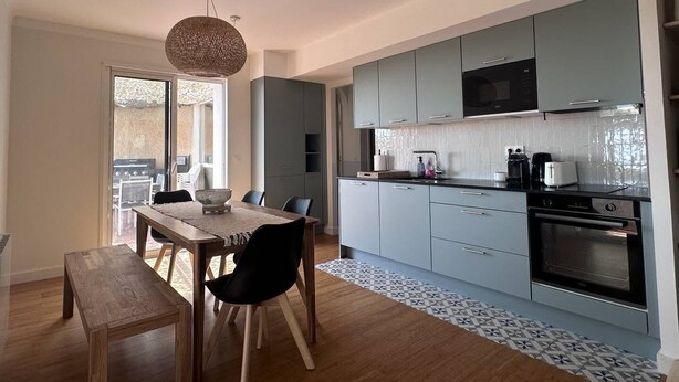 Une cuisine moderne avec des meubles gris, un four encastré et un coin repas comprenant une table et des chaises en bois.