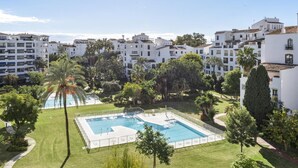 A pool area surrounded by white buildings, palm trees, and a clear blue sky. - Apartment in Terrazas de Banús for 4 people |4-12 (Puerto Banús - Marbella)