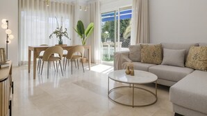 A modern living room with a sofa, coffee table, dining table, chairs, and a large window with curtains. - Apartment in Terrazas de Banús for 4 people |4-12 (Puerto Banús - Marbella)