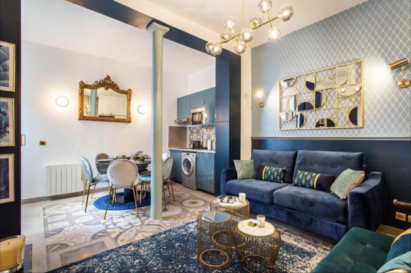 Un salon moderne avec un canapé bleu, un coin repas agrémenté d’un lustre et une cuisine équipée d’un four à micro-ondes.