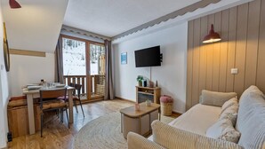 Un salon confortable avec un canapé, une table à manger et des chaises, un téléviseur à écran plat fixé au mur et une grande fenêtre donnant sur un paysage enneigé.