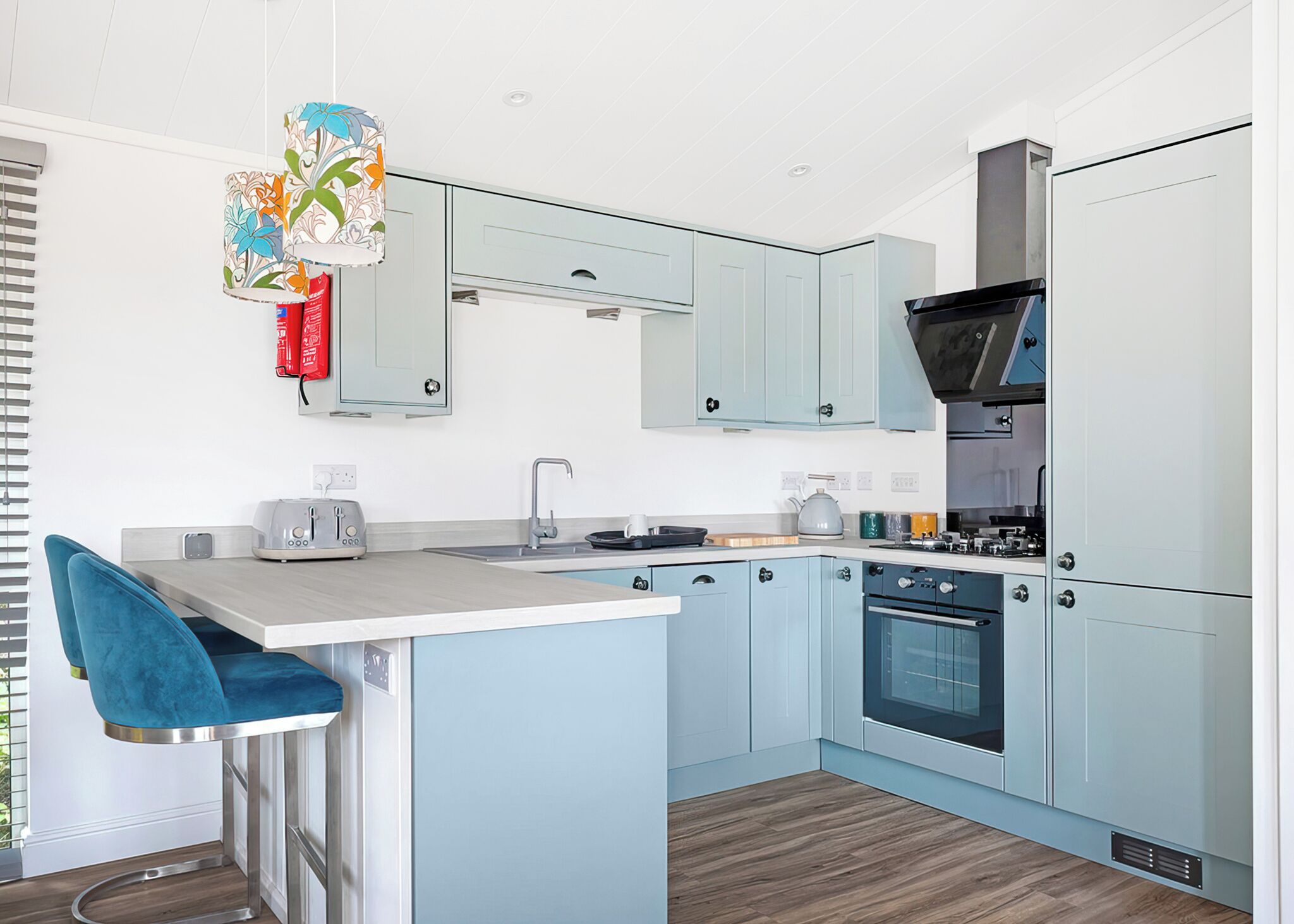 A modern kitchen with light blue cabinets, a central island with a bar stool, a built-in oven, and a hanging floral light fixture.