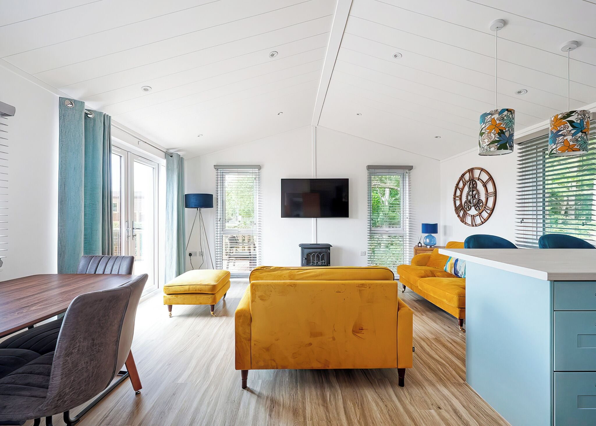 A modern living room with a dining area, a TV mounted on the wall, and a large window with blinds.