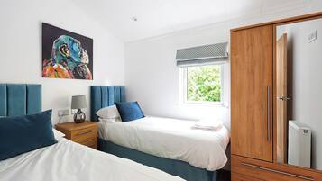 A bedroom with two beds, a wardrobe, a bedside table with a lamp, a window with a view of greenery, and a painting on the wall.
