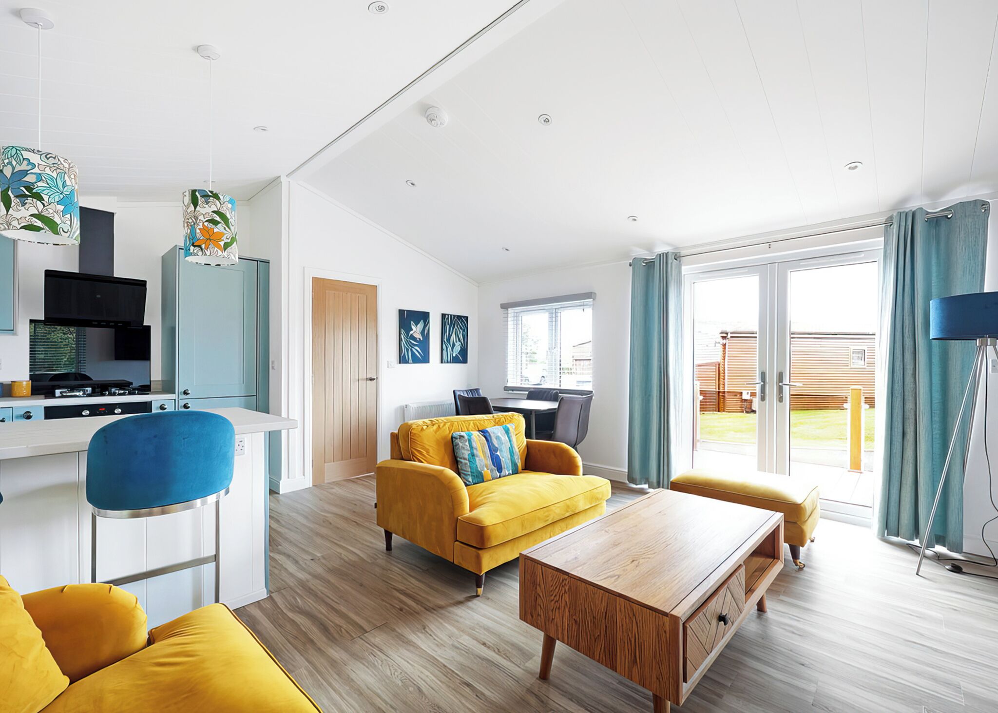 A modern living room with a yellow sofa, a wooden coffee table, and a kitchen area with a stove and cabinets.