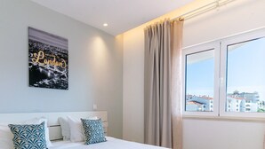 Ein Hotelzimmer mit Bett, einem Stadtbild-Poster, einem Fenster mit Vorhängen und Blick auf Gebäude.