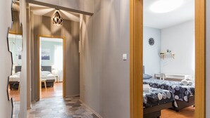 A hallway leading to a bedroom with a bed, a bedside table, and a nightstand. There is a mirror on the left wall, and a ceiling light fixture above the bed.