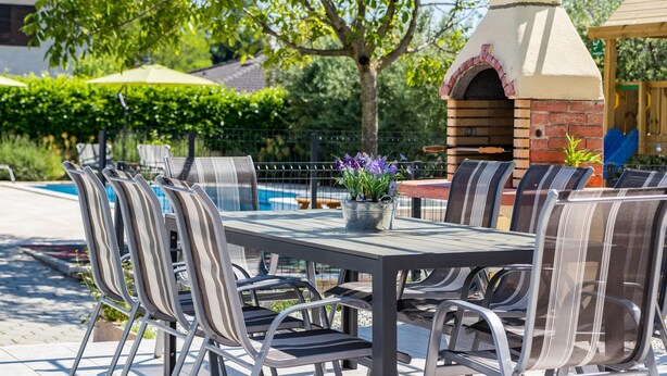 An outdoor patio with a brick pizza oven, metal chairs, and a table with a potted plant.