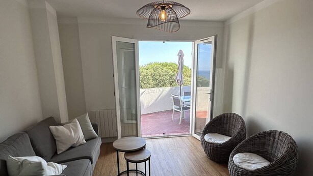 Ein modernes Wohnzimmer mit einem grauen Sofa, zwei Korbstühlen, einem kleinen runden Tisch und einer großen Schiebeglastür, die zu einer Außenpatio mit Tisch und Stühlen führt.