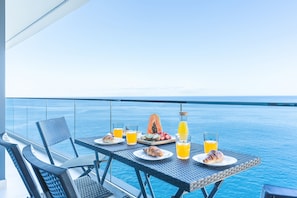 Une table à manger, dressée avec des aliments et des boissons pour le petit-déjeuner, sur un balcon donnant sur la mer.