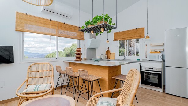 A modern kitchen with a central island, wicker chairs, and a large window offering a view of the outdoors.