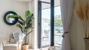 Una habitación con vistas a la costa, una silla, una planta en maceta, un espejo redondo y una mesita.