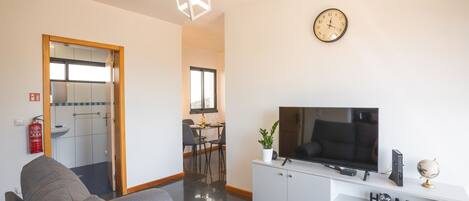 Una sala moderna con un sofá, una televisión, un mueble blanco para entretenimiento y un comedor al fondo.