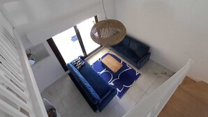 A high-ceiling room with a blue sofa set, a woven hanging light fixture, and a view through a large window.