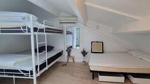 A dormitory room with bunk beds, a single bed, a small table with a lamp, a potted plant, and a mirror.
