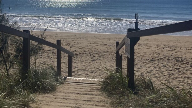 A wooden staircase leading to a beach with a clear view of the ocean and a sunny sky.