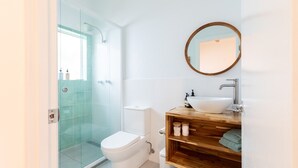 A modern bathroom with a glass shower enclosure, a white toilet, a wooden vanity with a sink, and a round mirror.