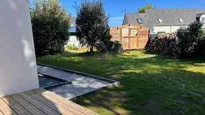 Eine Holzterrasse mit einer weißen, kugelförmigen Struktur, einem kleinen Wasserspiel und einem Gartenbereich mit einer Holzhütte und Bäumen.