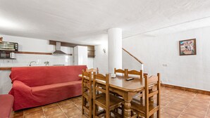 Een eetruimte met een houten tafel en stoelen, een rode bank, een keuken met witte tegels en een wit plafond.