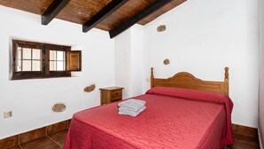 Een slaapkamer met een houten bed, een klein nachtkastje, een raam met houten kader en een betegelde vloer.
