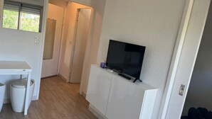 A modern room with a flat-screen TV on a white cabinet, a small table with a trash bin, and a door leading to another room. - 1 bedroom apartment with balcony near the beach (Saint-Raphaël)