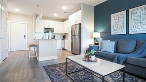 Una cocina moderna con armarios blancos y electrodomésticos de acero inoxidable, una isla central con taburetes y una sala de estar con sofá y arte abstracto en la pared.