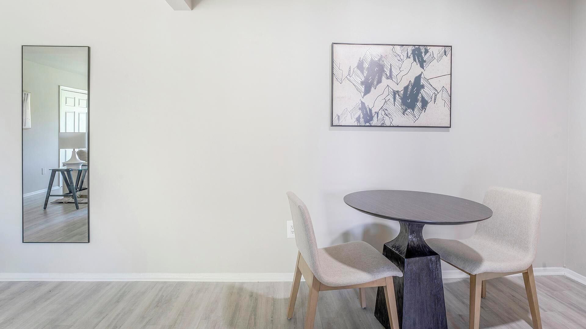 A dining area with a round table and two chairs, a large mirror, and a framed abstract artwork on the wall.