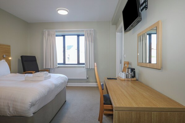 A hotel room with a bed, a desk, a chair, a TV, and a window with curtains. - Marine Court Suite (Littlehampton)