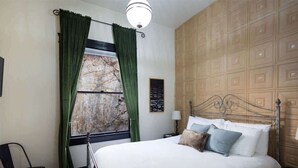 A bedroom with a large bed, a window with green curtains, a wooden headboard, and a wall-mounted light fixture. - The Block Basic Room - Queen (Bisbee)