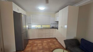 A compact kitchen with white cabinets, a refrigerator, and a window with blinds. - Comfortable Studio Granny Flat Mermaid Water (Mermaid Waters)
