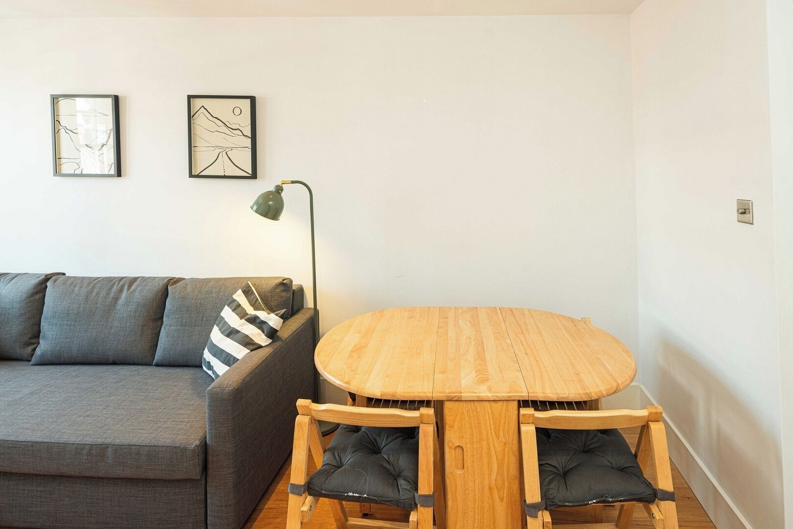 A small, modern living room with a grey sofa, a round wooden table with two chairs, and two framed artworks on the wall.