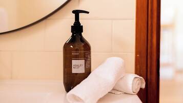 A bathroom with a bottle of Ashley & Co. hand soap and a rolled white towel on a white countertop.